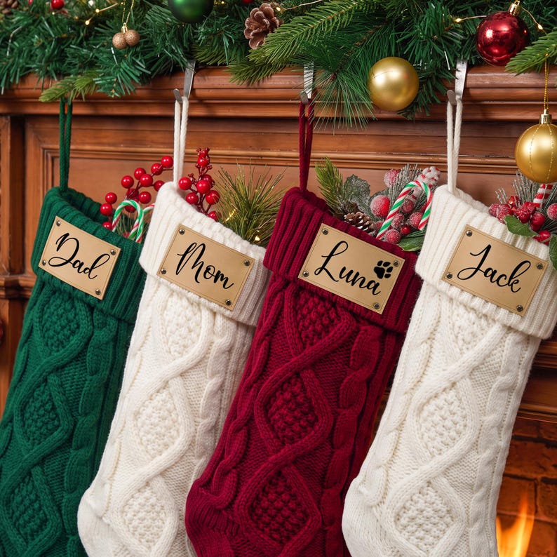May include: Four knit Christmas stockings in green, white, burgundy, and white. Each stocking features a cable knit pattern and a rectangular leather-like tag attached with metal rivets. The tags are personalized with the names Dad, Mom, Luna, and Jack.