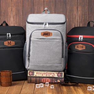 May include: Three insulated cooler backpacks in black and gray, with personalized leatherette patches. The black backpacks have a brown patch with a letter, while the gray backpack has a patch with the name "JAMES".