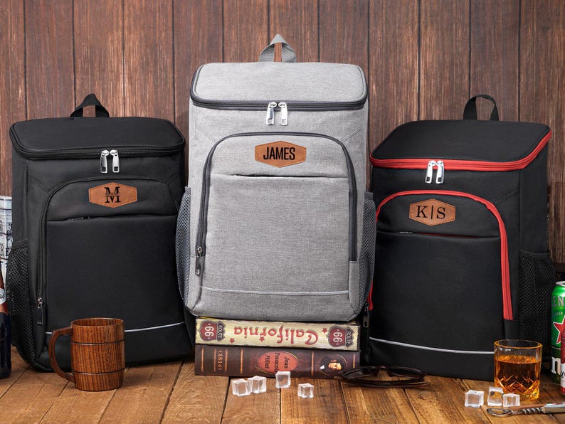 May include: Three insulated cooler backpacks in black and gray, with personalized leatherette patches. The black backpacks have a brown patch with a letter, while the gray backpack has a patch with the name "JAMES".