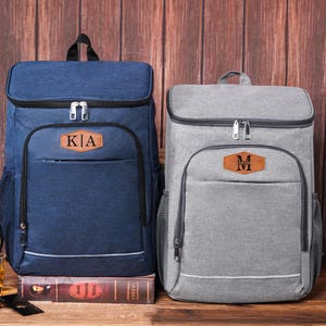 May include: Two insulated cooler backpacks, one navy blue and one gray, each with a front pocket and a leatherette patch with initials. The navy backpack has "KA" on the patch. The gray backpack has "M" on the patch. Both have a top handle and side mesh pockets.