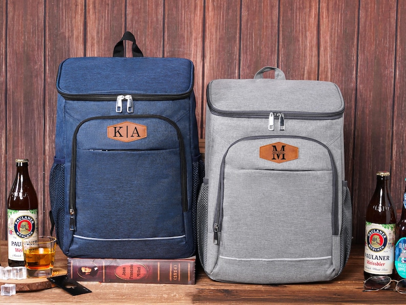 May include: Two insulated cooler backpacks, one navy blue and one gray, each with a front pocket and a leatherette patch with initials. The navy backpack has "KA" on the patch. The gray backpack has "M" on the patch. Both have a top handle and side mesh pockets.