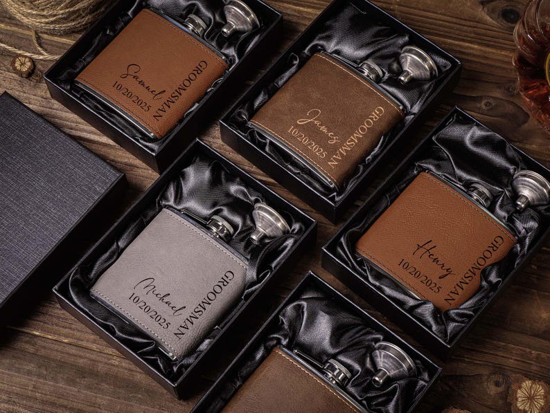 May include: Four brown leather-wrapped hip flasks with silver stoppers and personalised engravings. Each flask has the word "Groomisman" and a name, date, and the text "10.20.2025". The flasks are displayed in black boxes with a satin lining.