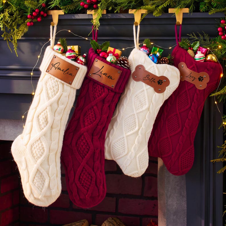 May include: Four cable-knit Christmas stockings in cream and deep red, each featuring a personalized leather patch. Two rectangular patches are engraved with the names Amelia and Liam, while two bone-shaped patches display the names Bella and Lucy.