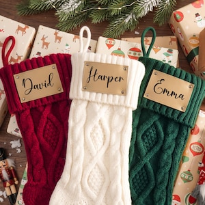 May include: Three cable-knit Christmas stockings in red, white, and dark green. Each stocking features a rectangular tan leather-like patch attached with four metal rivets. The patches are personalized with the names David, Harper, and Emma.