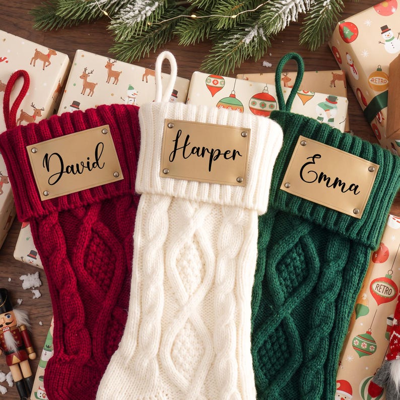 May include: Three cable-knit Christmas stockings in red, white, and dark green. Each stocking features a rectangular tan leather-like patch attached with four metal rivets. The patches are personalized with the names David, Harper, and Emma.