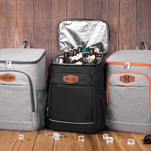 May include: Three personalized cooler backpacks in gray and black, with brown leatherette name tags. One black backpack is open, revealing bottles. The gray backpacks have orange trim. The backpacks are on a wooden surface.