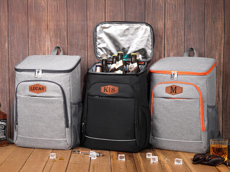 May include: Three personalized cooler backpacks in gray and black, with brown leatherette name tags. One black backpack is open, revealing bottles. The gray backpacks have orange trim. The backpacks are on a wooden surface.