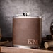 May include: A brown leather-wrapped stainless steel hip flask with a silver cap and the initials "KM" engraved on the front.