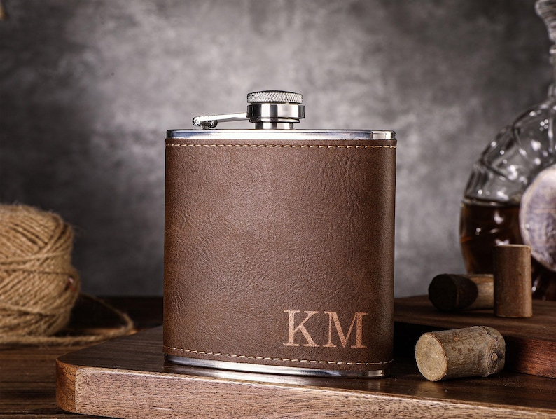 May include: A brown leather-wrapped stainless steel hip flask with a silver cap and the initials "KM" engraved on the front.