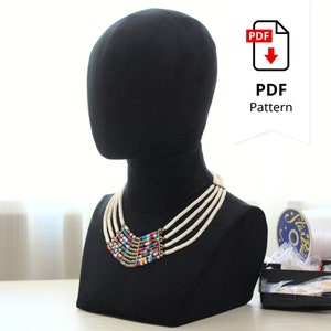 May include: A black velvet mannequin head displaying a multi-strand necklace. The necklace features white beaded strands and a central section with colorful beads. A PDF pattern icon is visible in the background.