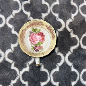 May include: A yellow teacup with a floral design featuring pink roses and green leaves. The cup has a gold rim and a white interior.