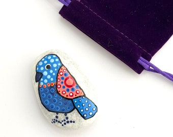 May include: Hand-painted rock with a blue, red, and white bird design. The rock is sitting on a white surface and is surrounded by a purple velvet bag.