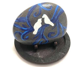 May include: A hand-painted stone with a blue and white design of two birds perched on a branch. The stone is displayed on a wooden stand.