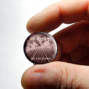 Puede incluir: Un cabujón redondo de vidrio con un diseño de esfera de reloj vintage. La esfera del reloj presenta números romanos y una vía de tren que conduce a una distancia brumosa. El cabujón se sostiene en una mano y tiene el texto "NIX CREATIONS" impreso en él.
