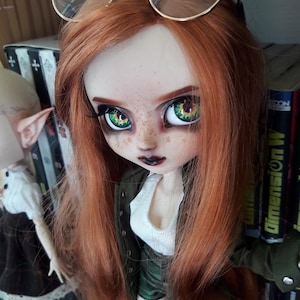 May include: A red-haired doll with green eyes and freckles wearing a green jacket, white shirt, and camouflage skirt. The doll is wearing glasses and has dark lipstick.
