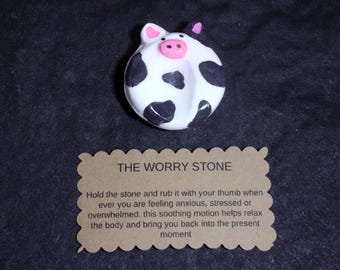 May include: A round white worry stone with black cow-like spots, pink ears, and a pink snout. Below it is a rectangular tan card with scalloped edges featuring the text: THE WORRY STONE. Hold the stone and rub it with your thumb when ever you are feeling anxious, stressed or overwhelmed. this soothing motion helps relax the body and bring you back into the present moment.