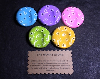 May include: Five circular worry stones in blue, purple, pink, green, and yellow, each featuring a white daisy pattern and a central thumb indentation. A rectangular brown card with scalloped edges displays the text THE WORRY STONE.