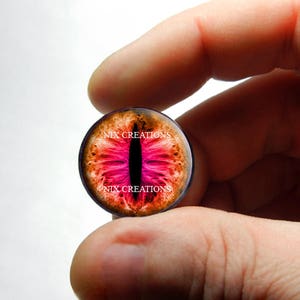 May include: Close-up of a round, glass cabochon with a pink and orange dragon eye design. The cabochon has the text "NIX CREATIONS" printed on it.