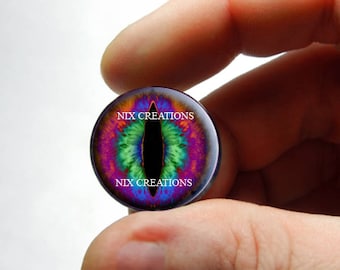 May include: A close-up of a colorful, iridescent dragon eye cabochon with a black pupil. The cabochon is set in a clear glass dome and has the text "NIX CREATIONS" printed on the top and bottom.