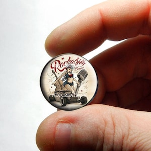 Pode incluir: Um cabochão redondo de vidro com uma ilustração vintage de uma mulher em uma pose de pin-up, sentada em um carro de corrida com um microfone e um violão. O texto "Rockabilly Boogie" está escrito em letras vermelhas e brancas na parte superior do cabochão.