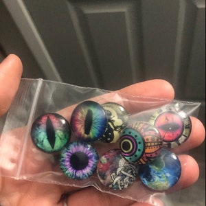 May include: A bag of colorful glass eyes with various designs, including a rainbow eye, a green eye with a black pupil, a purple eye with a black pupil, a black and white eye with a clock face, a colorful eye with a geometric design, and a blue and green eye with a galaxy design.