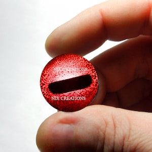 May include: Red glass eye with a black pupil and a textured surface. The eye has the text "NIX CREATIONS" printed on it.