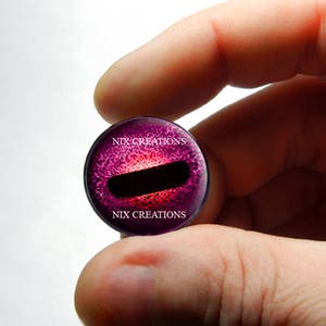 May include: A close-up of a purple and red glass eye with a black pupil. The eye is surrounded by a white border with the text "NIX CREATIONS" printed on it.