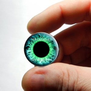 Op de afbeelding: Een groene en blauwe, oogvormige glazen cabochon met een zwarte pupil. De cabochon is omgeven door een zilverkleurige metalen rand. De tekst "NIX CREATIONS" is op de cabochon gedrukt.
