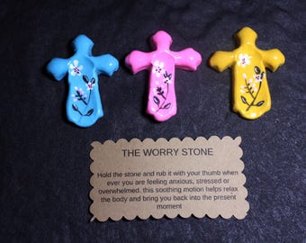 May include: Three cross-shaped worry stones in blue, pink, and yellow, each featuring a hand-painted white floral design. Below them is a scalloped-edge brown card with the text: THE WORRY STONE. Hold the stone and rub it with your thumb.