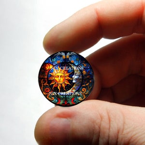 May include: A round glass cabochon with a stained glass design of the sun and moon. The sun is yellow with a human face and the moon is blue with a human face. The background is a dark blue with stars and swirls.