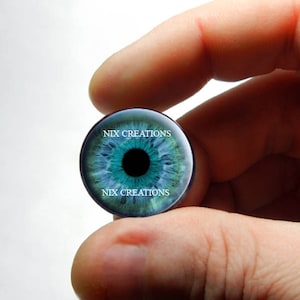 May include: A blue eye glass cabochon with a black pupil and white iris. The cabochon is held in a hand and has the text "NIX CREATIONS" printed on the top and bottom.