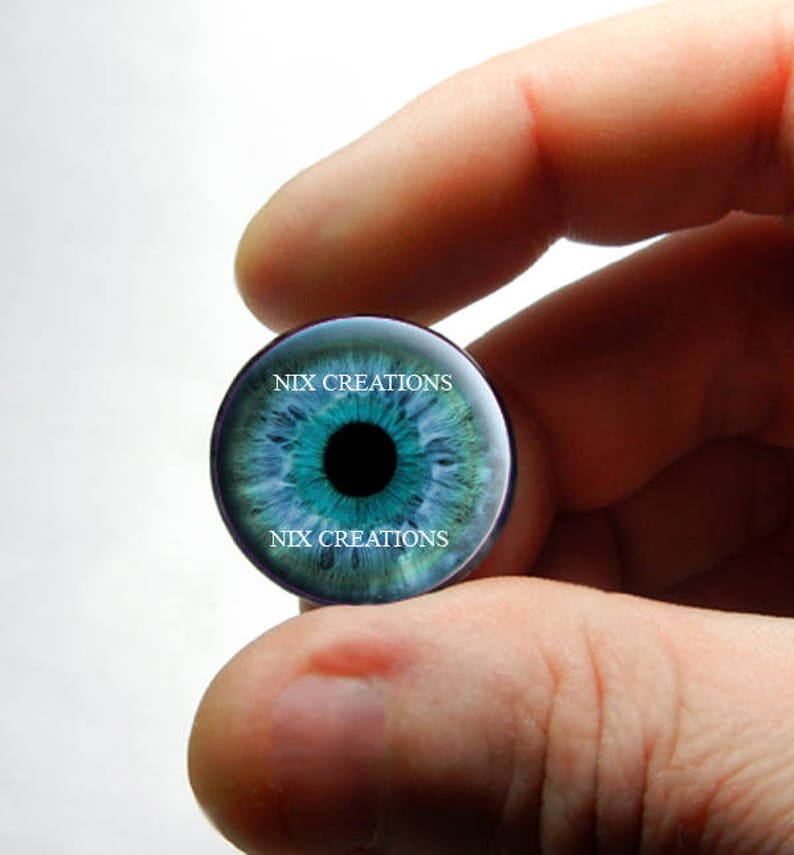 May include: A blue eye glass cabochon with a black pupil and white iris. The cabochon is held in a hand and has the text "NIX CREATIONS" printed on the top and bottom.