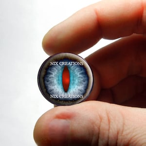 May include: A close-up of a round glass cabochon with a blue and white eye design and a red pupil. The cabochon is surrounded by a black border with the text "NIX CREATIONS" printed on it.