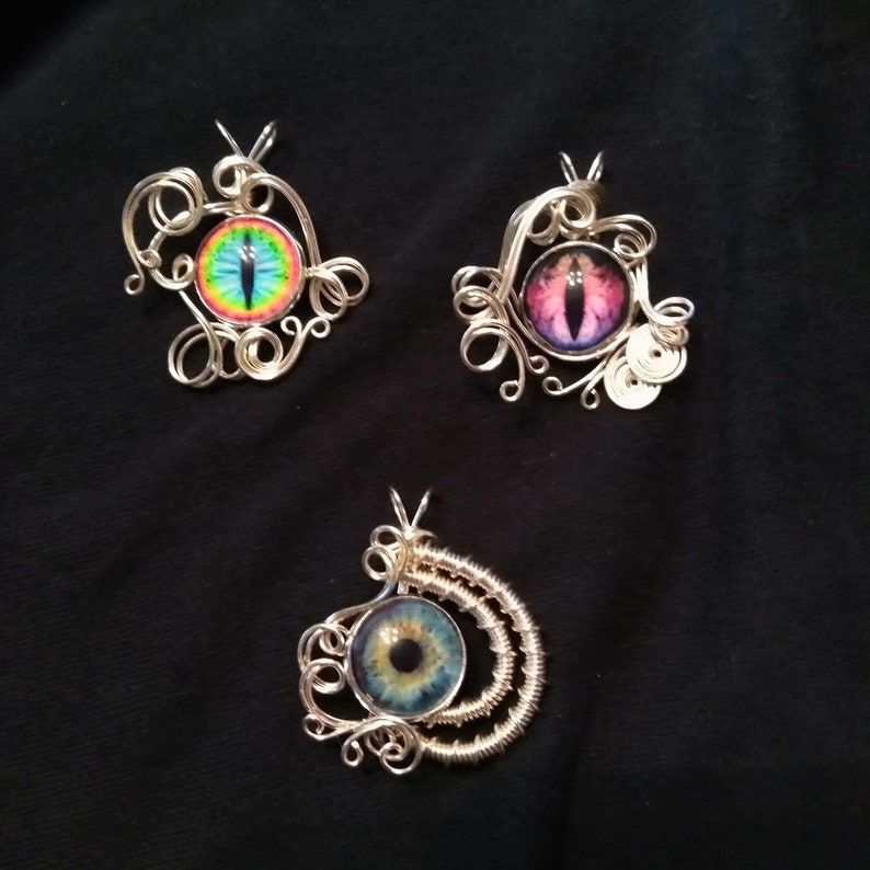 May include: Three silver wire wrapped pendants with colorful glass eyes. Each pendant features a different eye design with a blue, pink, and green color scheme.