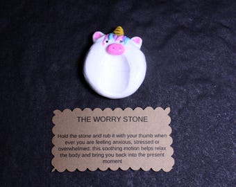 May include: A white, circular worry stone shaped like a unicorn with a gold horn, pink snout, and pastel-colored hair. Below it is a scalloped-edge brown card with the text: THE WORRY STONE. Hold the stone and rub it with your thumb when ever you are feeling anxious, stressed or overwhelmed. this soothing motion helps relax the body and bring you back into the present moment.