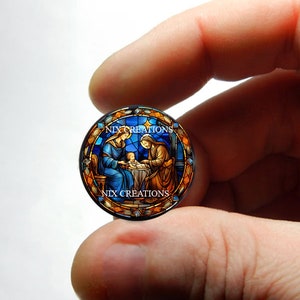 May include: A stained glass style round ornament featuring a depiction of the Holy Family, Mary, Joseph, and baby Jesus. The ornament is surrounded by a colorful border with the text "NIX CREATIONS" at the top and bottom.
