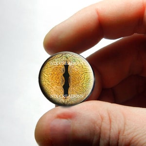 May include: A round, yellow and brown glass eye with a black pupil and a black, jagged line running down the center. The eye is surrounded by a black rim and the text "NIX CREATIONS" is printed on the top and bottom of the eye.