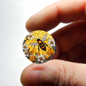 May include: A round, yellow, white, and black mosaic tile with a bee and flowers. The tile is surrounded by a black border and has the text "NYX CREATIONS" on the bottom.