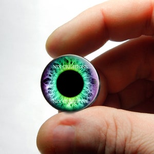 May include: A round glass cabochon with a colorful eye design. The eye is green, blue, and purple with a black pupil. The cabochon has the text "NIX CREATIONS" printed on it.
