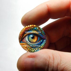 May include: A colourful eye with a blue iris and an orange pupil, surrounded by a mosaic of yellow, orange, blue, and green dots. The eye is painted on a round glass cabochon with the text "NIX CREATIONS" printed twice on the surface.