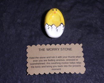 May include: A yellow and white painted stone shaped like a chick hatching from an egg, featuring a small indentation. Below the stone is a rectangular tan card with scalloped edges that reads: THE WORRY STONE. Hold the stone and rub it with your thumb.