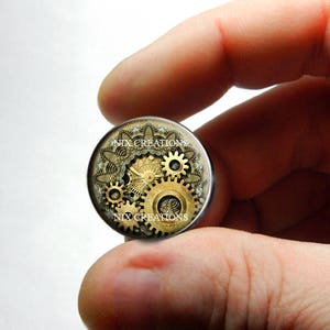 May include: A round, gold and black, steampunk-style, glass cabochon with a design of gears and a lace pattern. The cabochon has the text "NIX CREATIONS" printed on it.