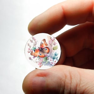 May include: A round glass cabochon with a painted image of a butterfly with orange and brown wings, surrounded by pink and white flowers.