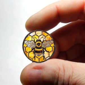 May include: A round glass cabochon with a bee and honeycomb design. The bee is yellow and black with brown wings. The honeycomb is yellow and brown.