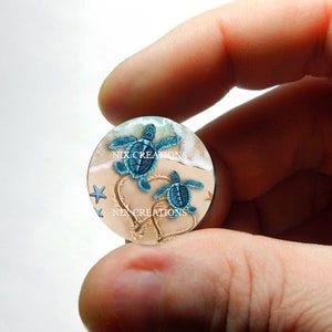 May include: A round, clear resin cabochon with a beach scene featuring two blue sea turtles swimming in the sand. The turtles are surrounded by white foam and blue water. The sand is imprinted with two heart shapes. The cabochon has a white background with the text "NIX CREATIONS" printed twice.