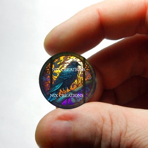 May include: A round stained glass piece with a black raven perched on a branch. The glass is iridescent and features shades of blue, purple, yellow, and orange. The text "NIX CREATIONS" is visible at the bottom of the piece.