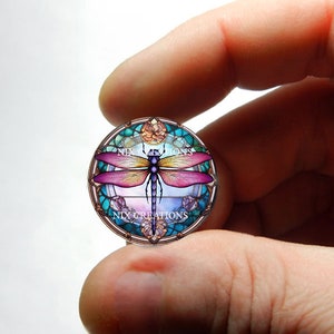 May include: A round glass cabochon with a stained glass design featuring a purple and yellow dragonfly with a blue body. The design is surrounded by a turquoise and gold frame with the text "Nix Creations".