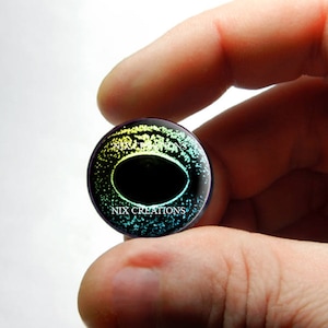 May include: A round, iridescent glass eye with a black pupil and a green, blue, and yellow ring around the pupil. The eye is surrounded by a black ring with the text "NIX CREATIONS" in white.