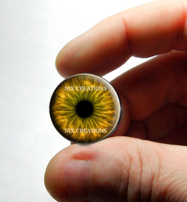 May include: A close-up of a yellow and black eye-shaped glass cabochon with the text "NIX CREATIONS" printed twice on the surface.