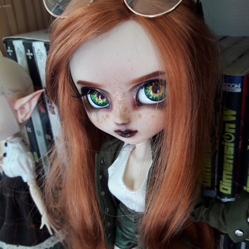 May include: A red-haired doll with green eyes and freckles wearing a green jacket, white shirt, and camouflage skirt. The doll is wearing glasses and has dark lipstick.